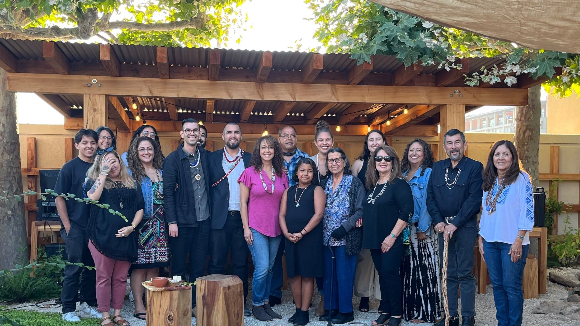 A large group of people pose for a photo under a wooden pavilion at an outdoor event, many wearing shell necklaces.