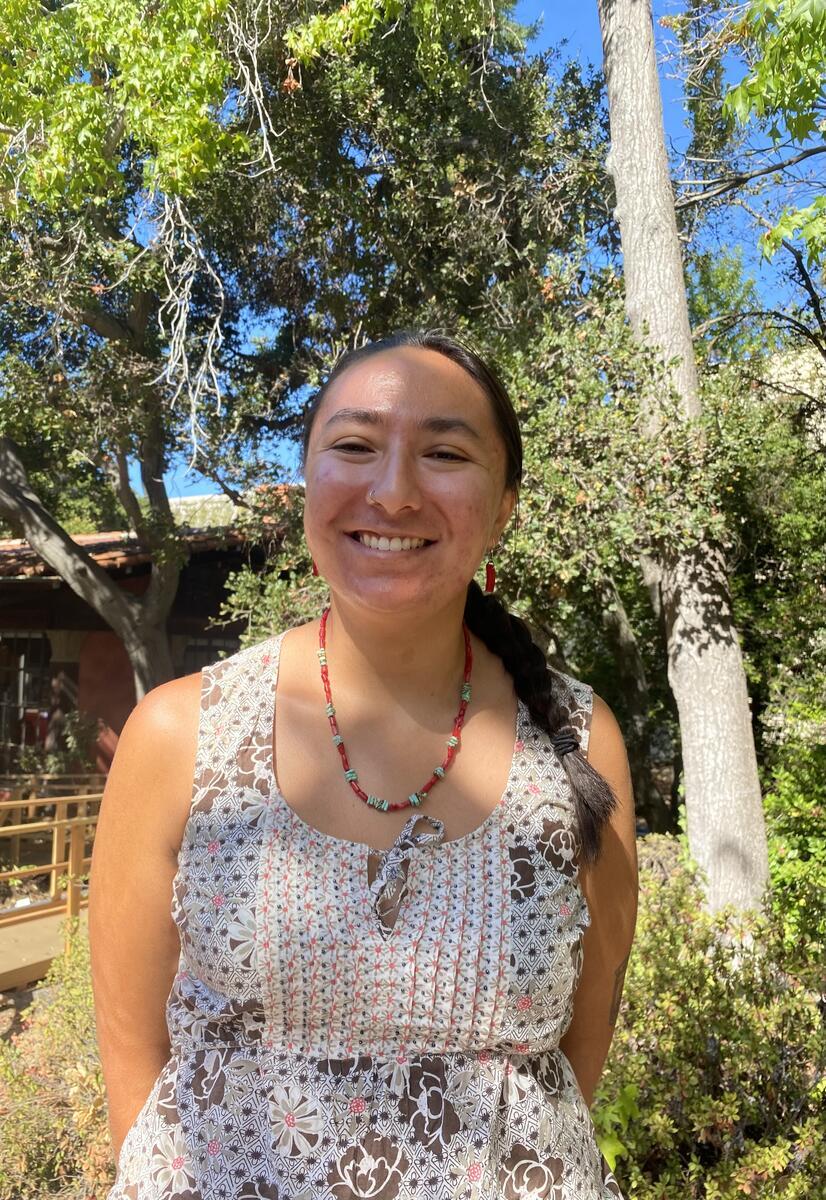 Headshot of Poppy Zingarelli-Gallegos wearing a patterned tank top and posed with large trees behind them.