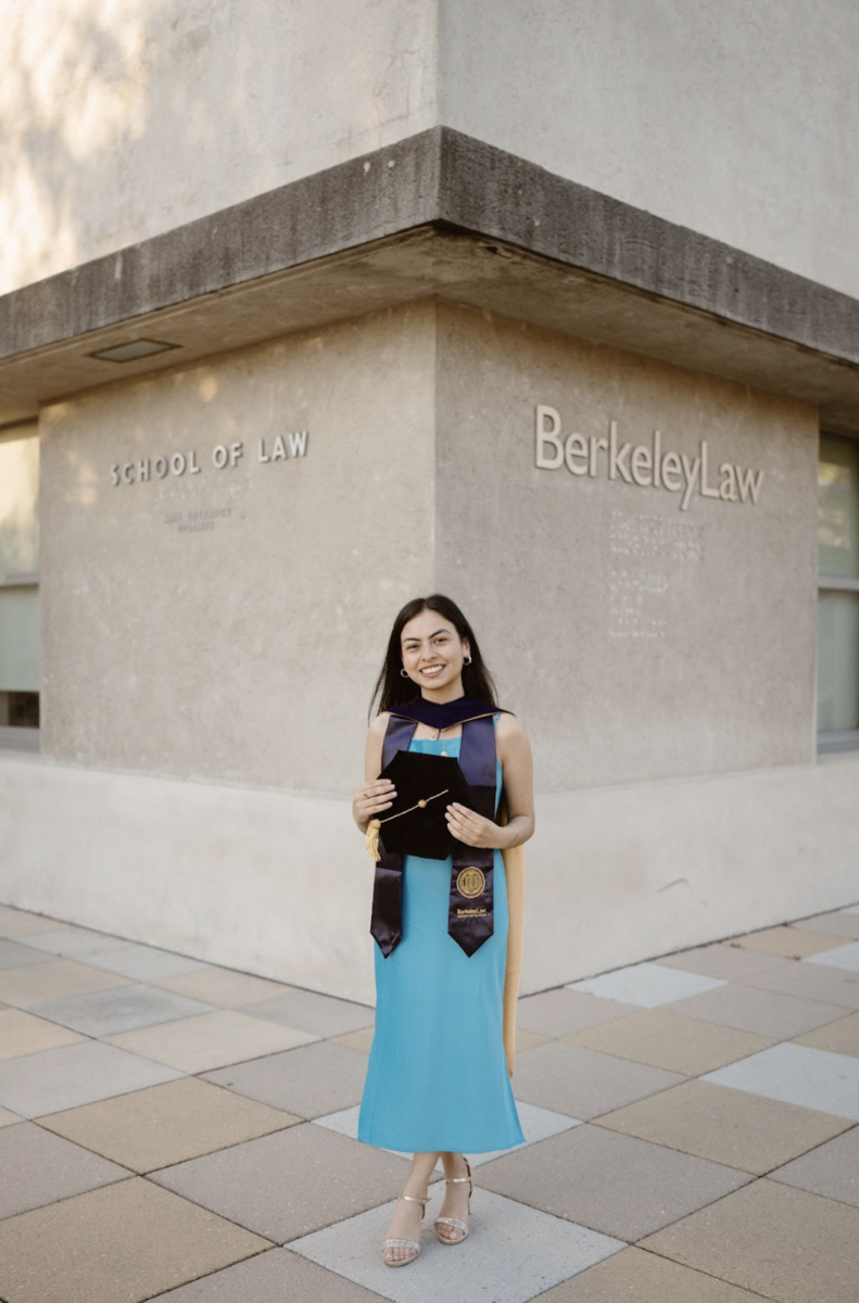 Angelica Rodriguez posed in front of Berkeley Law in a blue dress holding her mortarboard in front of her chest.
