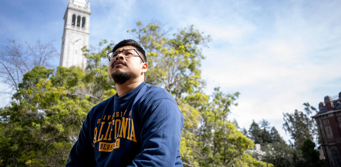 student looking up to the sky with the camera from a downwards angle looking up at the student with sather tower in the back